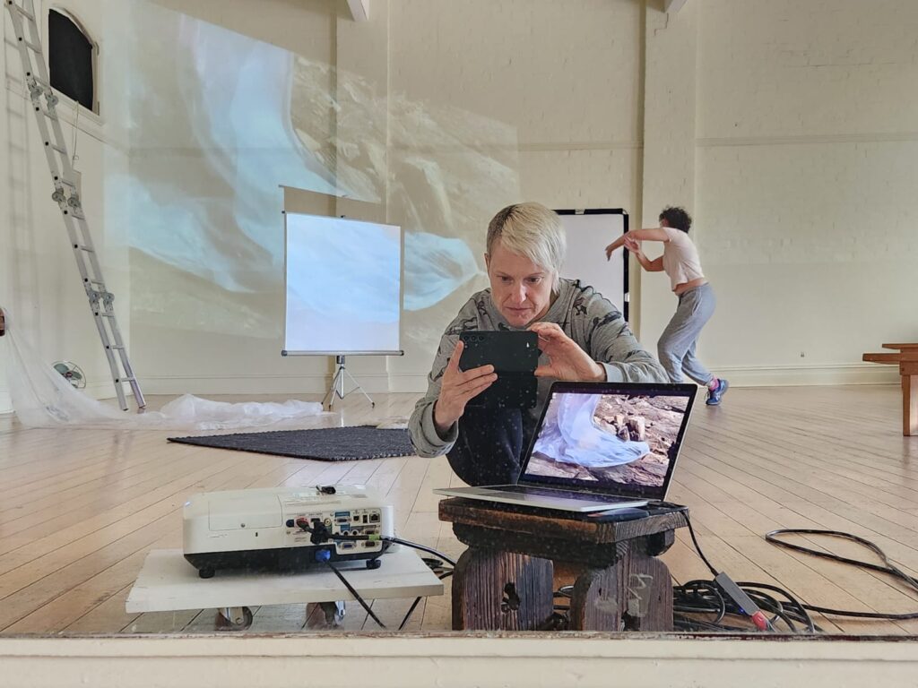 A person with short blonde hair takes a selfie in a mirror in front of them is a laptop behind them is another artist dancing.